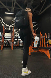 Person with a black backpack holding a white shaker cup in a gym setting.